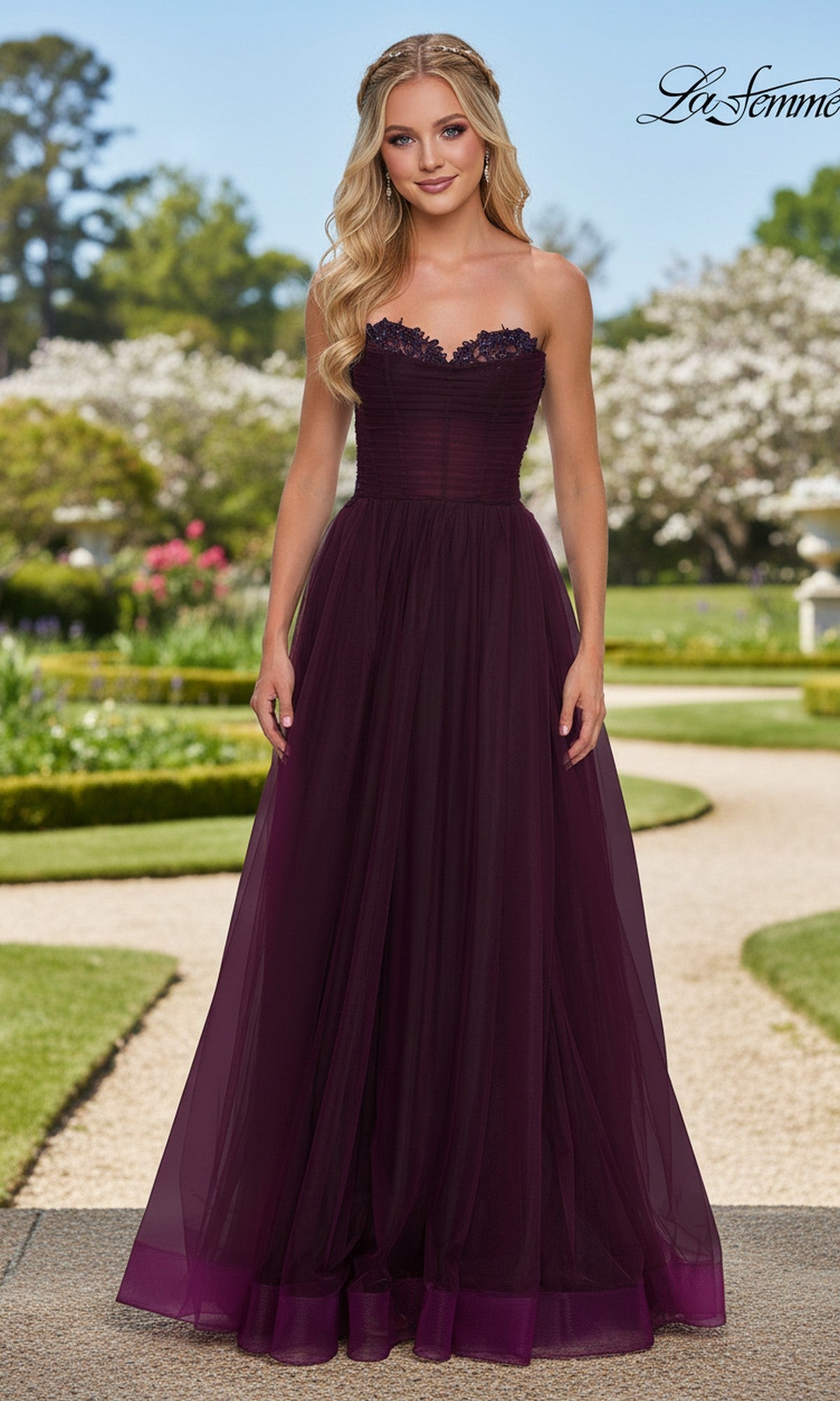 A woman poses elegantly outdoors in the La Femme 33363 long prom dress, a dark purple strapless gown with a fitted corset bodice and flowing tulle skirt, surrounded by greenery and blooming trees.